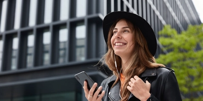 Photo charming dreamy woman holds modern cellular listens music walk outdoor metropolis