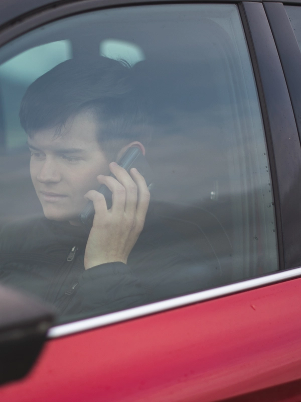 View young male wearing black jacket sitting red car while talking phone